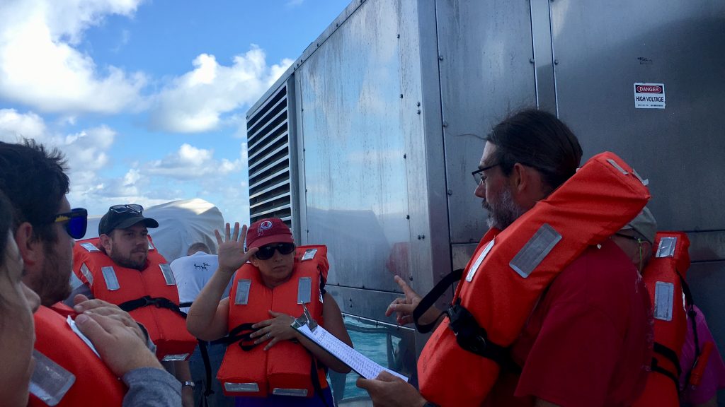 Michael holds a clipboard with the names of all participants aboard while 3 scientists surround him in life vests waiting for their name to be called.